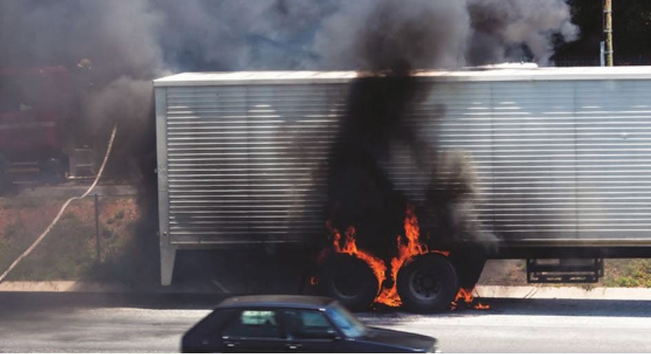 Truck wheels on fire on the side of a highway with firefighters on scene to extinguish the blaze.