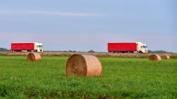 View of a field with hay in rolls against the background of trucks with semi-trailer driving along the highway. View of a field with hay in rolls against the background of trucks with semi-trailer driving along the highway.